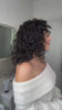Woman with dark curly shoulder-length hair turning sideways in a bathroom setting, wearing a white off-shoulder sweater, showcasing defined curls and natural hair texture.